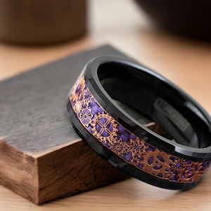 May include: A black ceramic ring with a rose gold and purple gear design. The ring is beveled and has a polished finish.