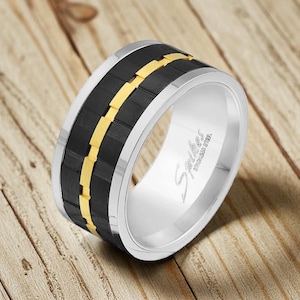 May include: A stainless steel ring with a black and gold design. The ring features a wide band with a black center section and a gold-colored stripe. The edges of the ring are silver. The ring has the word "Stainless Steel" engraved on the side.