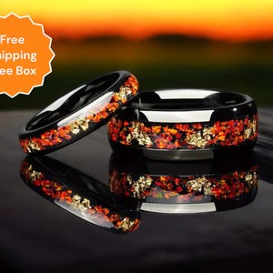 May include: Two black tungsten carbide rings with a red and gold inlay. The rings are on a black surface.