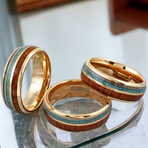 May include: Three gold-colored rings with a wood inlay and turquoise and white accents. The rings are displayed on a reflective surface, showcasing their polished finish and intricate design. The rings have a tungsten band.