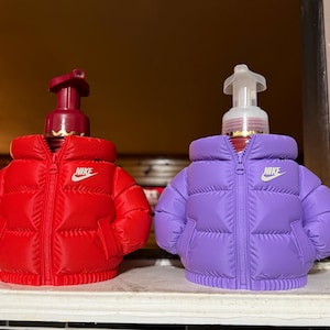 May include: Two soap dispensers shaped like puffer jackets, one red and one purple. Each has a white Nike logo on the chest. The dispensers have a pump top and a zipper detail.