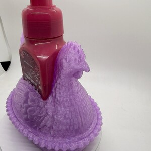 May include: A decorative, lavender-colored chicken-shaped soap dispenser holder. The holder cradles a dark pink soap bottle with a pump. The soap bottle has a label that reads "Strawberry". The holder is made of a textured material.