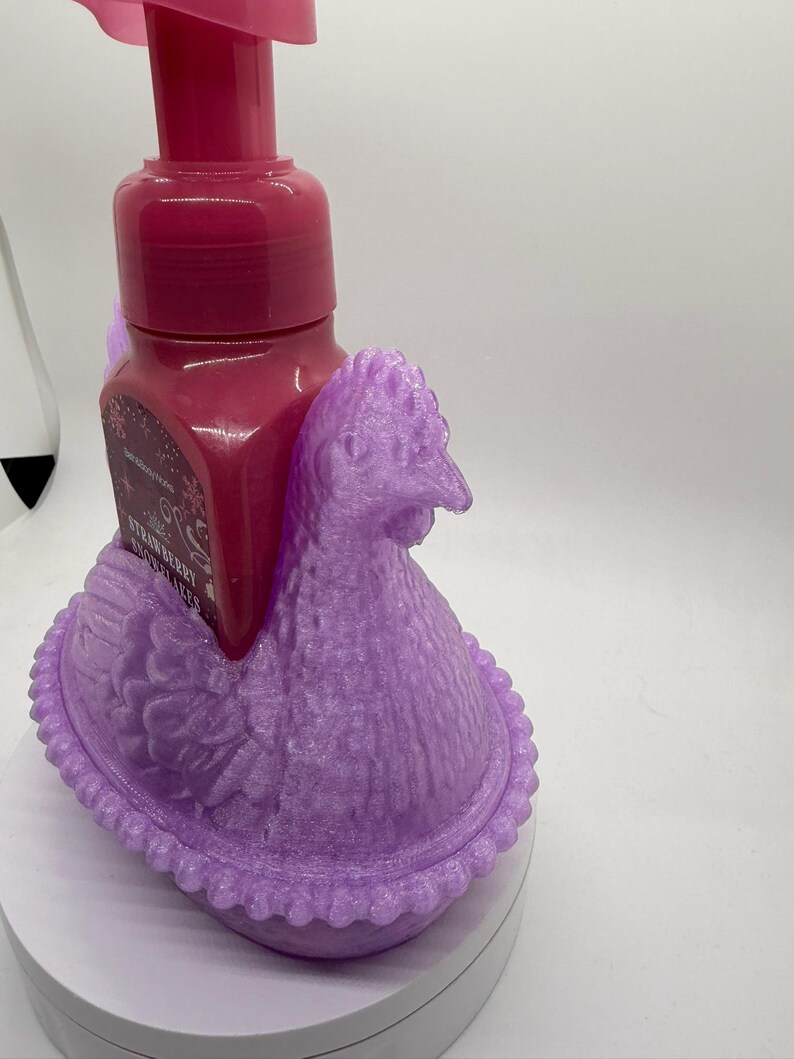 May include: A decorative, lavender-colored chicken-shaped soap dispenser holder. The holder cradles a dark pink soap bottle with a pump. The soap bottle has a label that reads "Strawberry". The holder is made of a textured material.