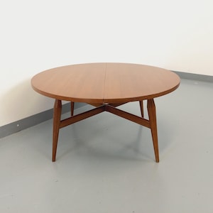 May include: A round wooden table with a drop leaf extension. The table has a brown finish and a simple, mid-century modern design.