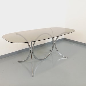 May include: Oval-shaped dining table with a smoked glass top and a chrome base. The base features a series of curved, interconnected supports. The table's dimensions are approximately 60 inches long and 36 inches wide.