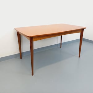 Op de afbeelding: Een rechthoekige houten eettafel met een lichtbruin tafelblad en vier taps toelopende poten. De tafel is ongeveer 152 cm lang en 76 cm breed, met een eenvoudig, modern design uit het midden van de eeuw. De tafel staat tegen een witte muur en een grijze vloer.