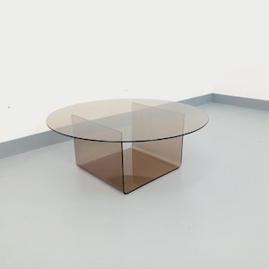 May include: A modern, round coffee table with a transparent, brown-tinted glass top. The base is a geometric design, also in a brown hue. The table's design is minimalist and contemporary, suitable for living room decor.