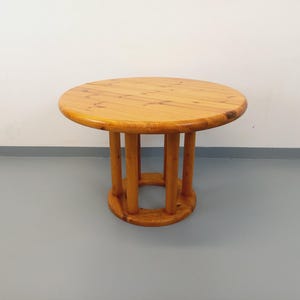 May include: A circular wooden table in a light brown hue. The table features a round top supported by vertical wooden posts connected to a base. The table is made of wood and displays a natural wood grain pattern.