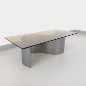 May include: A modern coffee table with a rectangular, tinted glass top. The table has a curved, brushed metal base. The table's dimensions are approximately 48 inches long, 24 inches wide, and 16 inches high.