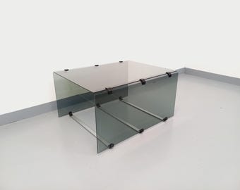 May include: A smoked glass coffee table with a rectangular top and a lower shelf. The table is supported by a metal frame with black clips.