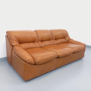 Könnte beinhalten: Ein Dreisitzer-Sofa aus braunem Leder. Das Sofa hat ein niedriges Profil, abgerundete Armlehnen und eine getuftete Rückenlehne. Die Kissen wirken weich und einladend. Geeignet für ein Wohnzimmer oder einen Loungebereich.