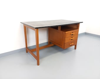 May include: A mid-century modern desk with a rectangular top and three drawers. The desk has a dark gray top with a black border, supported by wooden legs and a wooden drawer unit. The drawers have small, rounded wooden handles.