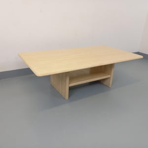 May include: A light-colored rectangular coffee table with a lower shelf. The table has a simple, modern design with a smooth surface and rounded corners. The table is made of wood and is approximately 48 inches long and 24 inches wide.