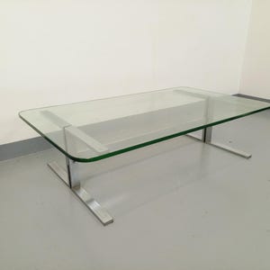 May include: A rectangular glass coffee table with a polished metal base. The clear glass top has rounded corners and is supported by two metal bars. The base consists of two rectangular metal supports. The table is in a room with white walls and a gray floor.