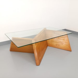 May include: A mid-century modern coffee table with a clear glass top and a wooden base. The base is constructed of light-colored wood and features a unique geometric design. The table is rectangular and sits on a gray floor.