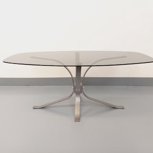 May include: A smoked glass coffee table with a silver metal base. The base has four legs that curve out and then back in, creating a unique design.