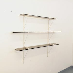 May include: Three-tiered wall-mounted shelving unit. The shelves are made of dark tinted glass, supported by white metal brackets. The unit is mounted on a white wall, providing a minimalist and modern aesthetic. The shelves are evenly spaced.