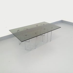 Op de afbeelding: Een rechthoekige salontafel met een gerookte glazen bovenkant en een chromen metalen onderstel. De tafel heeft een minimalistisch ontwerp. De tafel is ongeveer 122 cm lang en 61 cm breed. Het glas heeft een grijze tint.