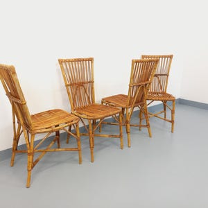 May include: Four vintage bamboo chairs with woven seats and backs. The chairs have a natural, light brown color and a classic design. The chairs are arranged in a row against a white wall and a gray floor.