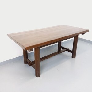 Pode incluir: Uma mesa de jantar retangular de madeira com um acabamento liso em castanho médio. A mesa apresenta um tampo espesso e sólido suportado por quatro pernas cilíndricas robustas ligadas por uma barra transversal horizontal entre as pernas.