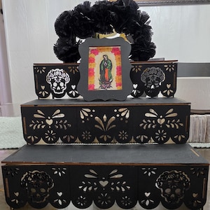 May include: A three-tiered black wooden display stand featuring intricate laser-cut patterns of skulls and floral motifs. A small black frame containing an image of the Virgin of Guadalupe surrounded by yellow and pink flowers sits on the top tier.