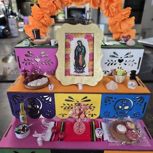 May include: A colorful Day of the Dead altar with a gold-framed image of the Virgin Mary, surrounded by miniature food items, flowers, and decorative boxes. The altar is adorned with orange paper flowers and a traditional Mexican table runner.