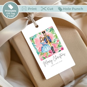 Könnte beinhalten: Ein weißer Geschenkanhänger mit einer Aquarell-Illustration eines Nussknackers und einer Ballerina vor rosa Blumen. Der Text "Merry Christmas" und "Happy New Year" ist in schwarzer Kursivschrift geschrieben.