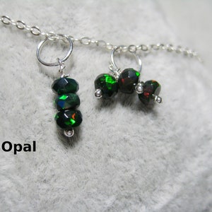 Könnte beinhalten: Eine silberne Kettenkette mit zwei Anhängern. Ein Anhänger hat drei grüne Opalperlen und der andere Anhänger hat drei schwarze Opalperlen. Das Wort "Opal" steht unter den Anhängern.
