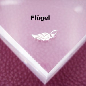 Könnte beinhalten: Ein silberner Anhänger in Form eines Flügels. Der Anhänger befindet sich auf einem weißen Hintergrund mit dem Wort "Flügel" darüber.