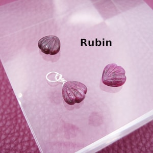 Könnte beinhalten: Drei rosafarbene herzförmige Rubin-Steine, einer an einer silbernen Kette, auf einer weißen Oberfläche. Das Wort "Rubin" ist in schwarzer Schrift auf der Oberfläche geschrieben.