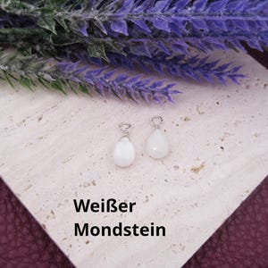 May include: Two white teardrop-shaped pendants with silver-colored metal loops. The pendants are on a light beige surface, with purple flowers in the background. The text "Weißer Mondstein" is below the pendants.