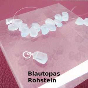 May include: A collection of light blue aquamarine rough stones on a clear surface. The stones are arranged in a line and are of varying sizes. The text "Blautopas Rohstein" is printed below the stones.