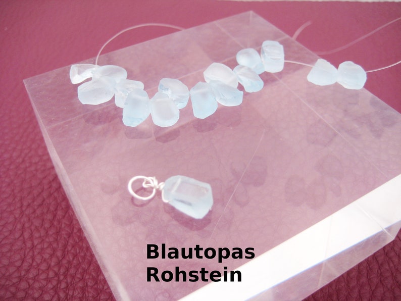 May include: A collection of light blue aquamarine rough stones on a clear surface. The stones are arranged in a line and are of varying sizes. The text "Blautopas Rohstein" is printed below the stones.