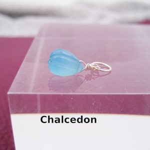 May include: A light blue chalcedony gemstone pendant with a silver wire wrap. The pendant is on a pink surface with the word "Chalcedon" written below it.