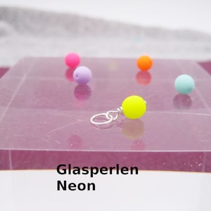 Può includere: Primo piano di una perla di vetro di colore giallo neon con un anello di salto argentato. La perla è su una superficie rosa con altre perle di colore neon sullo sfondo. Il testo "Glasperlen Neon" è visibile nella parte inferiore dell'immagine.