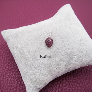 Op de afbeelding: Een klein, rood robijn hanger in de vorm van een blad, op een wit fluwelen kussen. Het woord "Rubin" staat onder de hanger.