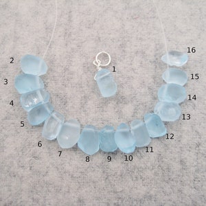 May include: A necklace made of 16 light blue sea glass beads. The beads are irregular in shape and have a frosted finish. The necklace is strung on a clear cord and has a silver charm attached.