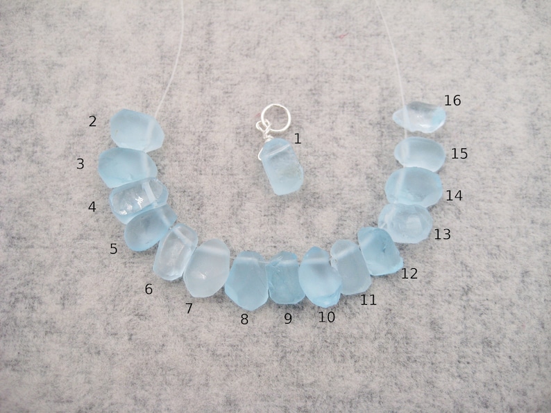 May include: A necklace made of 16 light blue sea glass beads. The beads are irregular in shape and have a frosted finish. The necklace is strung on a clear cord and has a silver charm attached.