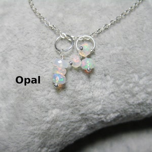 Könnte beinhalten: Eine silberne Kettenkette mit einem Cluster aus kleinen weißen Opalperlen, die daran hängen. Die Kette liegt auf einer weißen, flauschigen Oberfläche. Das Wort "Opal" ist in schwarzer Schrift unterhalb der Kette geschrieben.