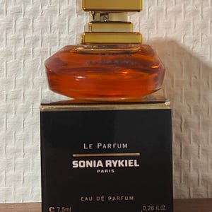 Może przedstawiać: Miniaturowy szklany flakon perfum z bursztynowym płynem i złotą zakrętką, umieszczony na czarnym pudełku z napisem Le Parfum Sonia Rykiel Paris Eau de Parfum, 7,5 ml, 0,26 fl. oz. Flakon posiada szeroką, zaokrągloną podstawę i kwadratową górę.