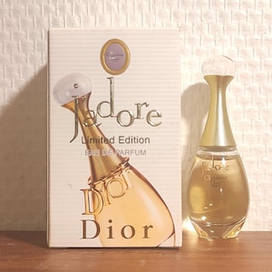 May include: A bottle of "J'adore" by Christian Dior perfume, alongside its box. The perfume bottle has a gold neck and a clear, rounded top. The box is white with the perfume bottle design and the words "J'adore" and "Dior".