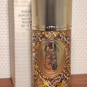 Puede incluir: Un frasco de perfume con una tapa de metal dorado y un cuerpo de vidrio decorativo. El cuerpo presenta un estampado floral en tonos dorados y marrones. La etiqueta dice "MADAME ROCHAS" y "PARFUM DE TOILETTE".