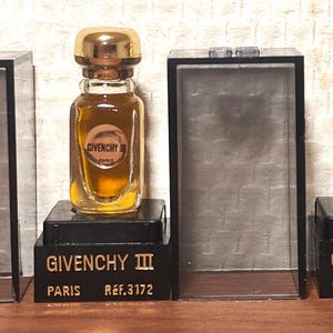 May include: Three vintage Givenchy III perfume bottles with gold caps and amber liquid, displayed on black stands. Each stand has the Givenchy III logo and "Paris Ref. 3172" printed on it. The bottles are in clear display cases.