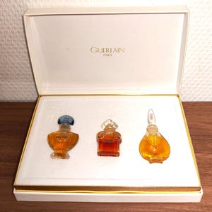 Könnte beinhalten: Eine weiße Präsentationsbox mit dem goldenen Schriftzug GUERLAIN PARIS im Deckel. Die Box enthält drei Miniatur-Parfümflakons aus Glas mit bernsteinfarbener Flüssigkeit. Die Flakons sind mit SHALIMAR, L'HEURE BLEUE und einem dritten beschriftet.