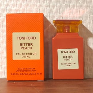 May include: A Tom Ford Bitter Peach eau de parfum 7.5 ml bottle and its matching orange box. The box features the text Tom Ford Bitter Peach Eau de Parfum 7.5 ml and 0.25 fl. oz./oz. liq. 7.5 ml e. The bottle has a peach-colored body and a yellow cap.