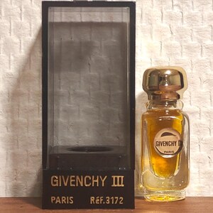 May include: A miniature perfume bottle with a gold cap and a clear glass body, filled with a golden liquid. The bottle is next to a black display case with the words "GIVENCHY III PARIS REF.3172" in gold lettering.