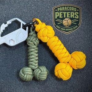 May include: A multi-tool key chain with a silver metal key-shaped tool and two paracord keychains. One keychain is olive green, and the other is yellow. The key chain has the text "PARACORD PETERS" and "KNOT YOUR AVERAGE GEAR."