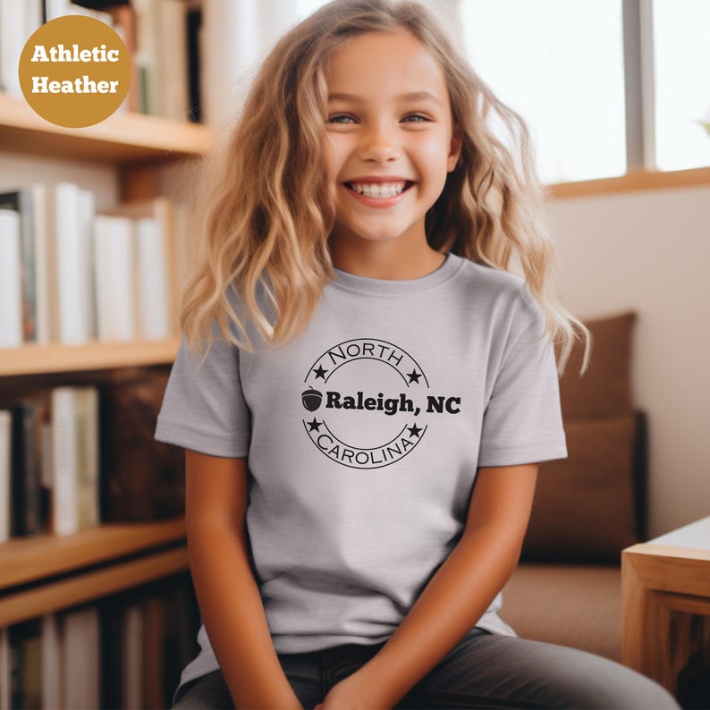 May include: A young person wearing a light gray t-shirt with a black and white graphic of a circle with the text "NORTH CAROLINA" and a small acorn graphic. The text inside the circle reads "Raleigh, NC".