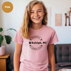 May include: A pink t-shirt with a black circle graphic that says "NORTH CAROLINA" with a star on each side and an acorn graphic above the text. The text inside the circle says "Raleigh, NC".