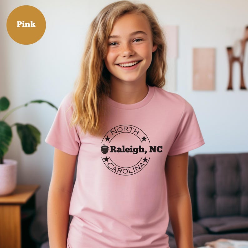 May include: A pink t-shirt with a black circle graphic that says "NORTH CAROLINA" with a star on each side and an acorn graphic above the text. The text inside the circle says "Raleigh, NC".